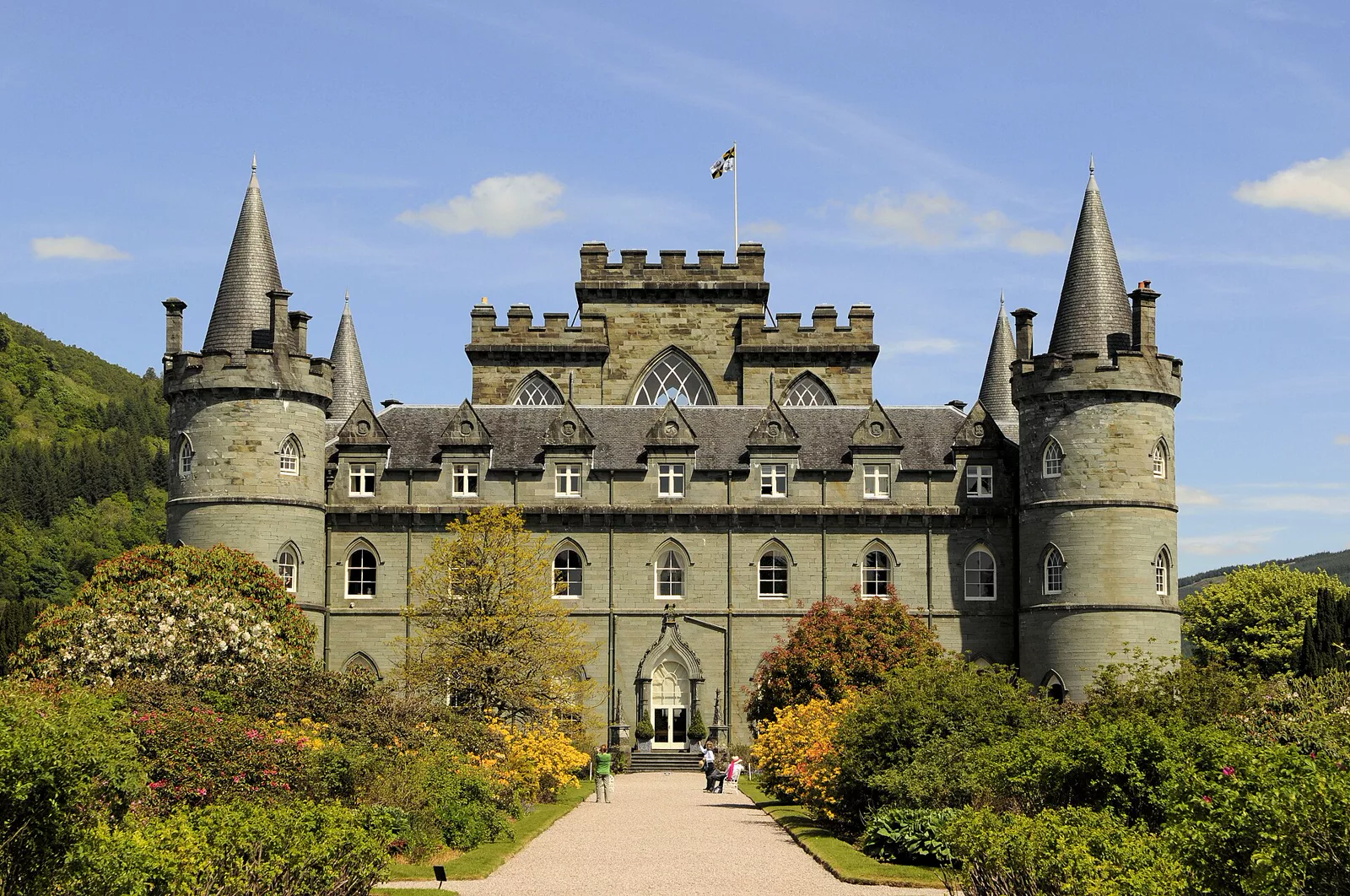 Inveraray Castle, a grey stone castle with four round corner towers with conical roofs, seen along a path through garden shrubs under a blue sky.