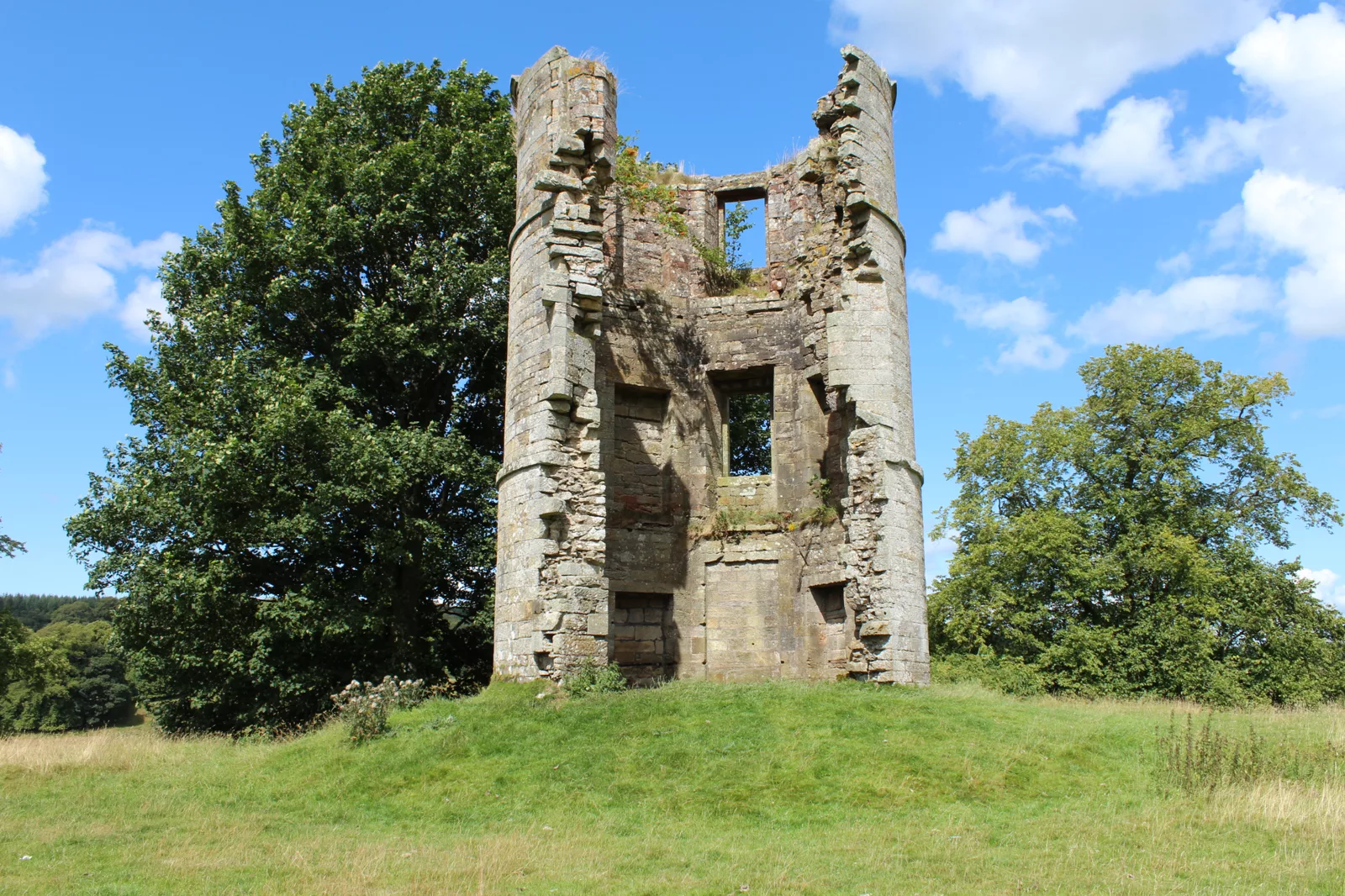 The remains of a round-cornered stone tower on a grassy mound, with trees on either side and a blue sky behind.