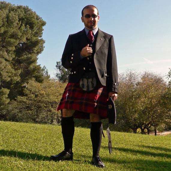 A man standing outdoors in a red and dark tartan kilt with a black jacket, sporran and black kilt hose.