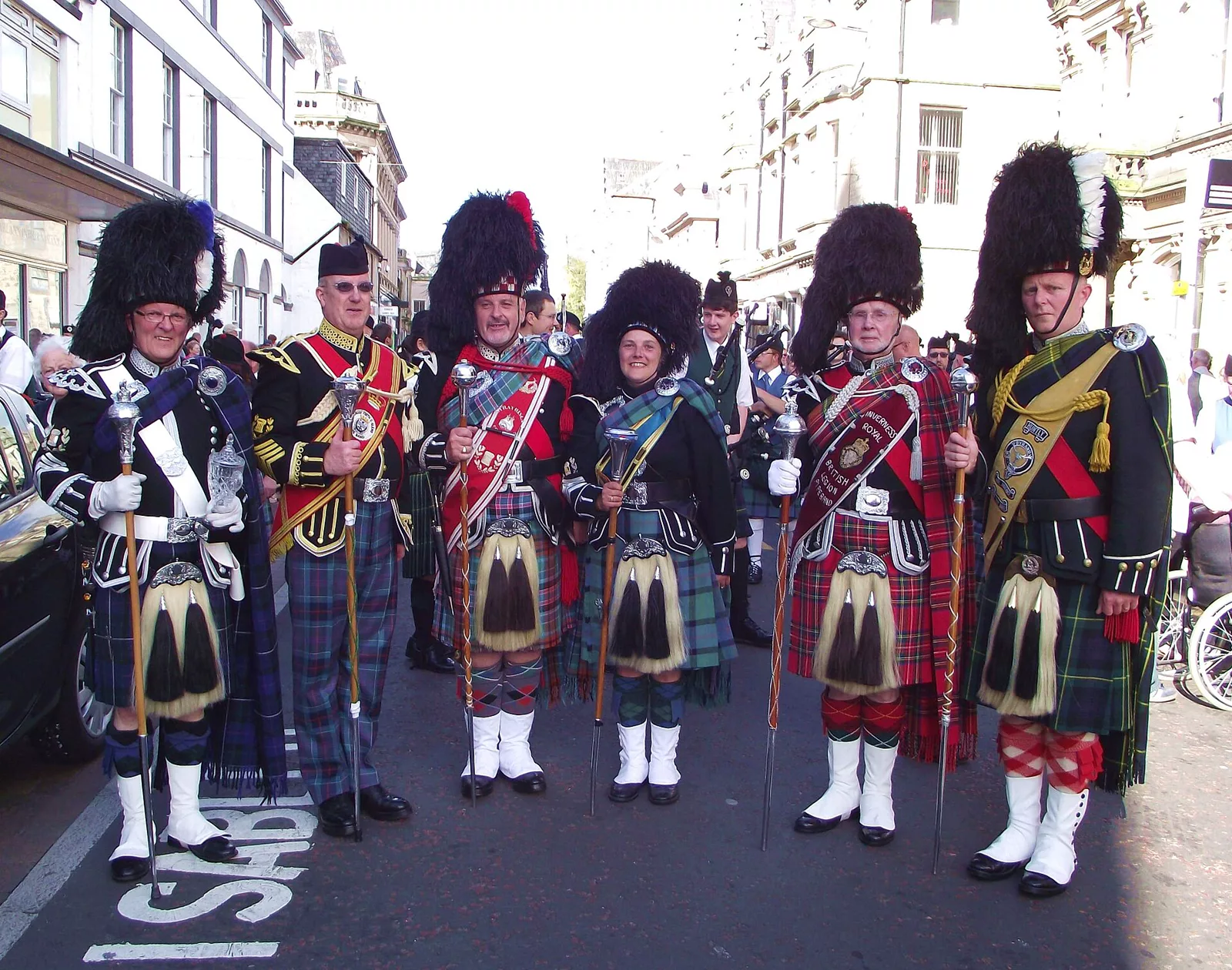 Six pipe band drum majors in full Highland dress, with kilts, sporrans, sashes and feather bonnets, standing together in a town street