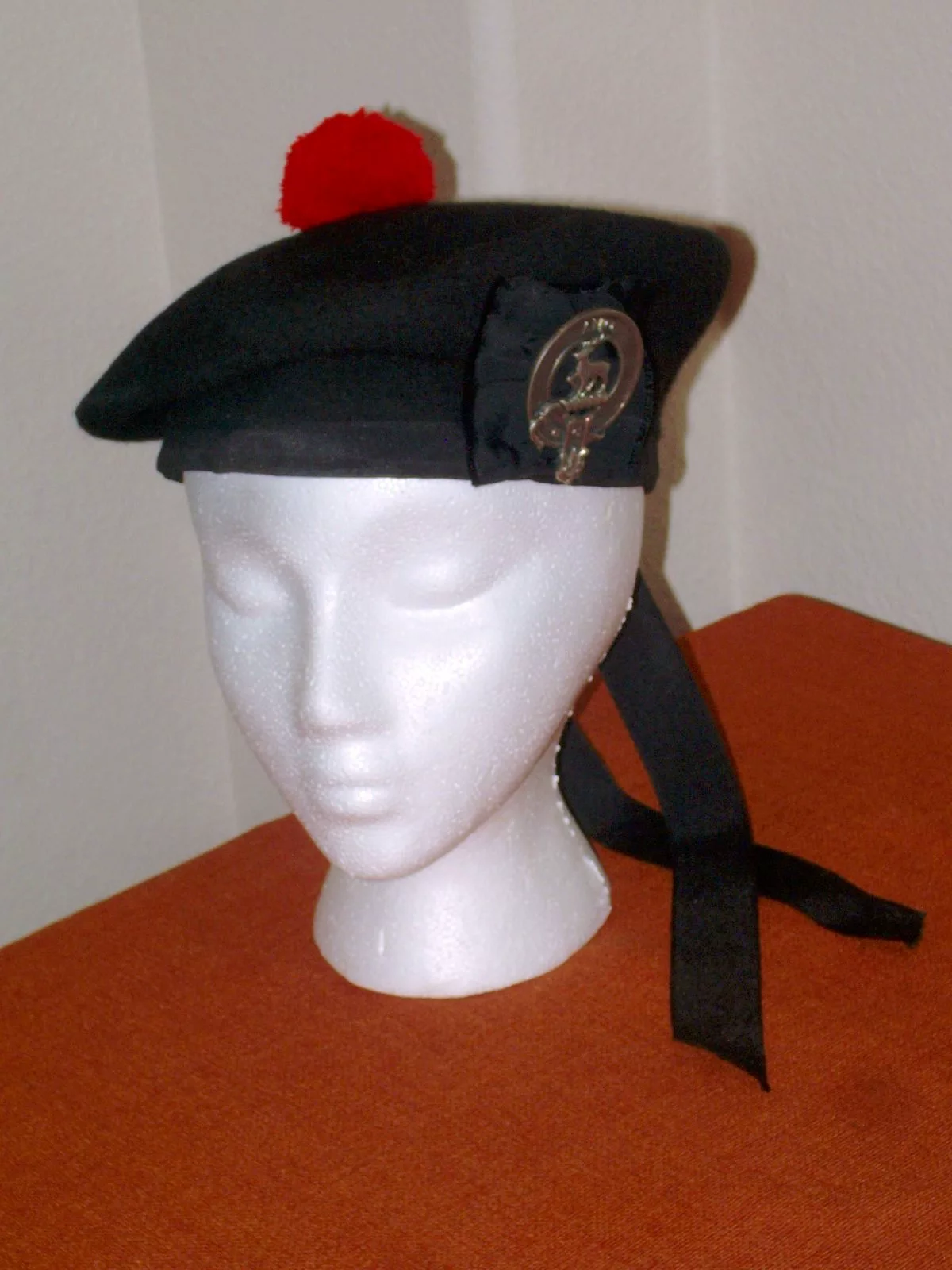 A black Balmoral bonnet with a red toorie, a clan crest badge and ribbon tails, shown on a white foam head.