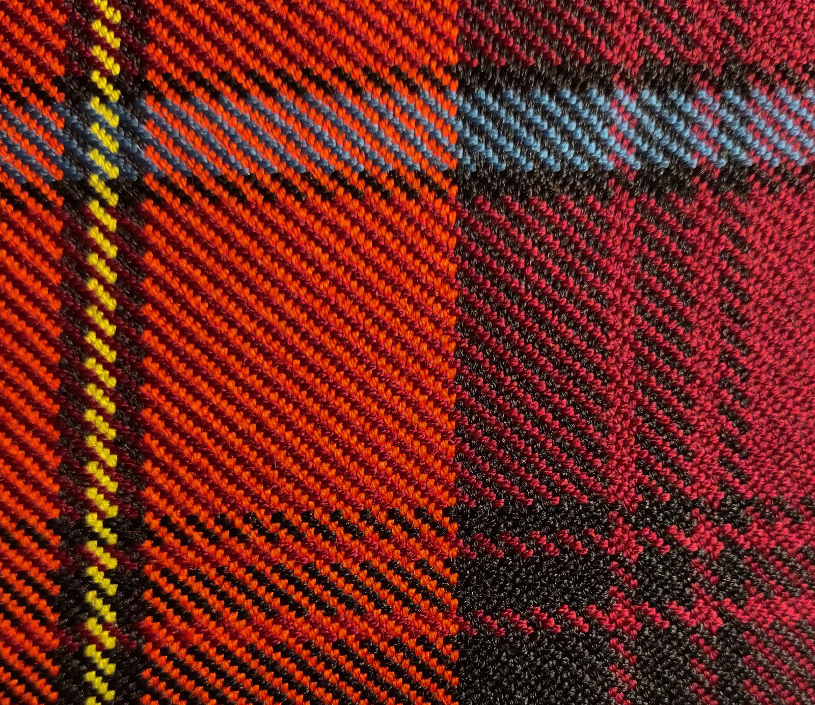 Close-up of woven tartan cloth showing the diagonal twill ribs in red, crimson, blue, yellow and black yarns.