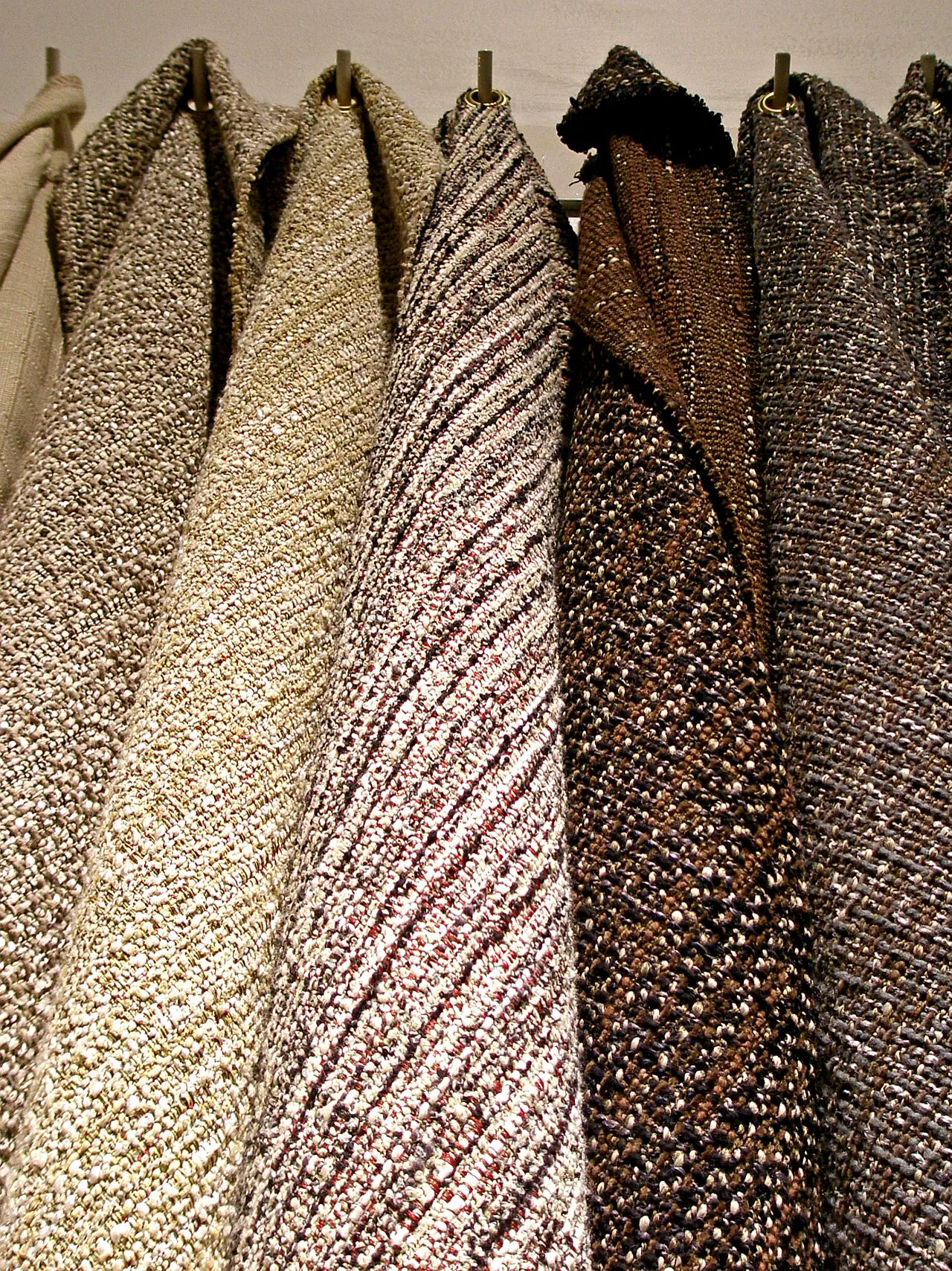 Several woven tweed cloths in cream, brown, maroon and grey hanging from hooks, each with a different texture and weave.